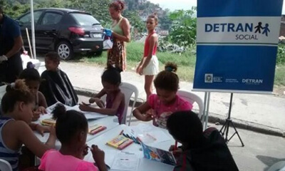 Em uma comunidade do Rio de Janeiro, seis crianças estão colorindo desenhos numa mesa em estrutura montada pelo Detran. Ao fundo uma menina, ao lado de sua mãe, observa as outras crianças.