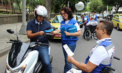 Dois agentes educativos do Detran, uma mulher de óculos e um cadeirante, orientam um motociclista que também recebe brindes. Ao fundo outros agentes fazem o mesmo com um taxista.