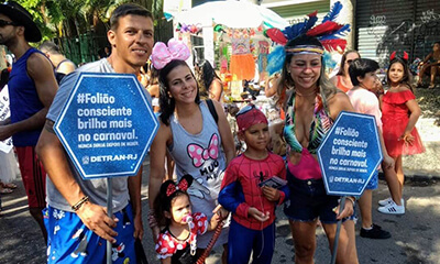 Três adultos e duas crianças, sendo o menino fantasiado de homem-aranha e a menina de Minnie, pulam carnaval em bloco de rua. Ao fundo há mais pessoas fantasiadas.
