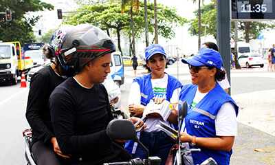 Duas funcionárias do Detran orientam motociclistas na orla de Copacabana, que também recebem brindes.