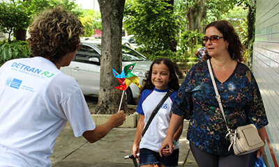 Funcionária do Detran entrega catavento a menina sorridente que anda de mãos dadas com sua mãe a caminho da escola.