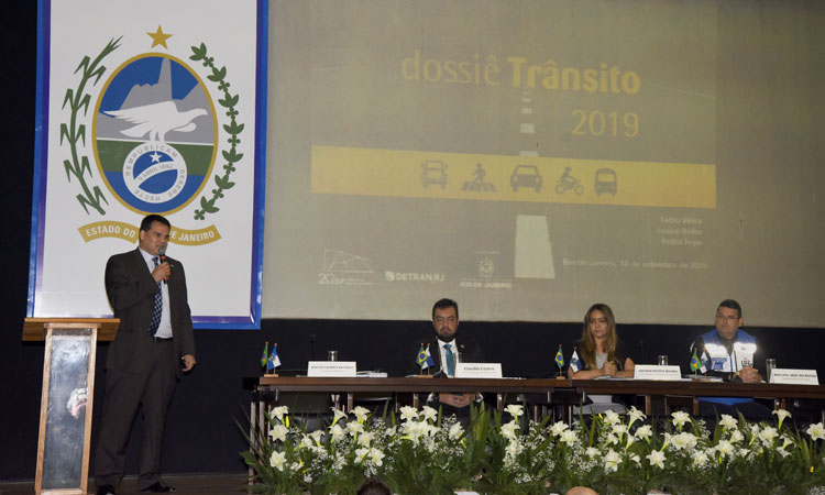 Presidente do Detran discursa no palco acompanhado de mais 3 participantes do debate sobre Dossiê do Trânsito. Ao fundo um grande banner com a imagem do brasão do Estado do Rio de Janeiro