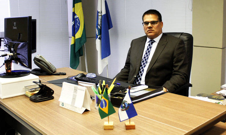 Presidente do Detran, vestindo terno, em seu gabinete de trabalho composto por 2 bandeiras grandes ao fundo, uma do Brasil e outra do Estado do Rio de Janeiro