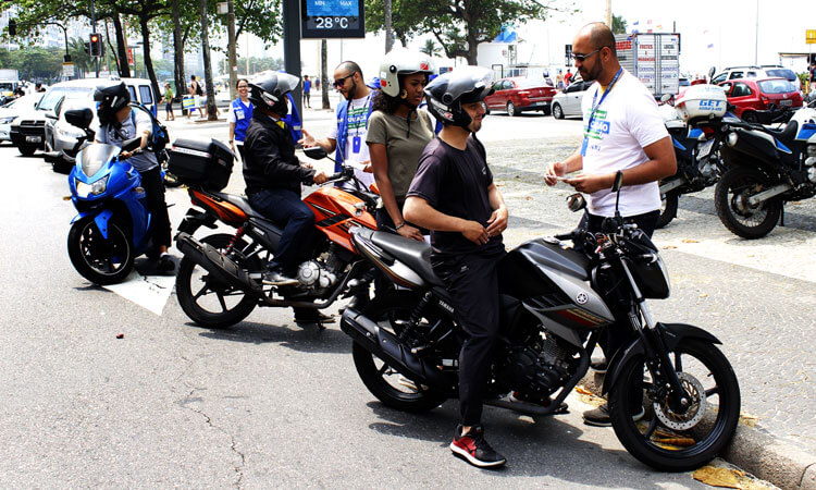 Dois agentes do Detran passam orientações a dois motociclistas em acostamento próximo à orla de Copacabana. Ao fundo um relógio/termômetro de rua marca 28 graus
