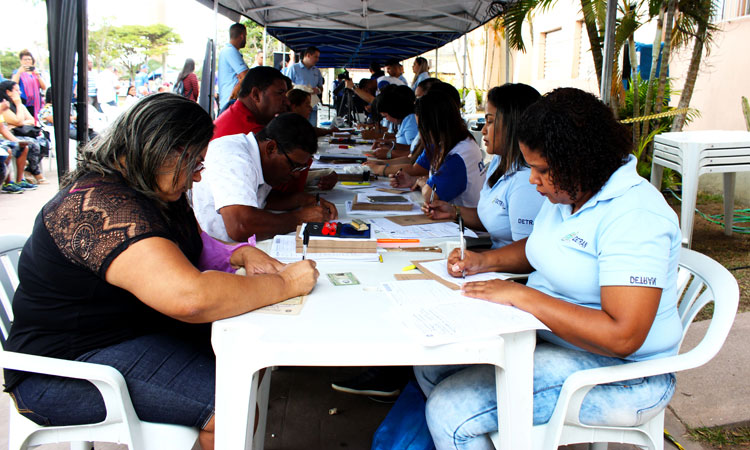 Embaixo de toldos e sentados diante de funcionários do Detran, cidadãos cariocas preenchem formulário para solicitar a emissão da carteira de identidade. Outras pessoas aguardam sentadas a sua vez.
